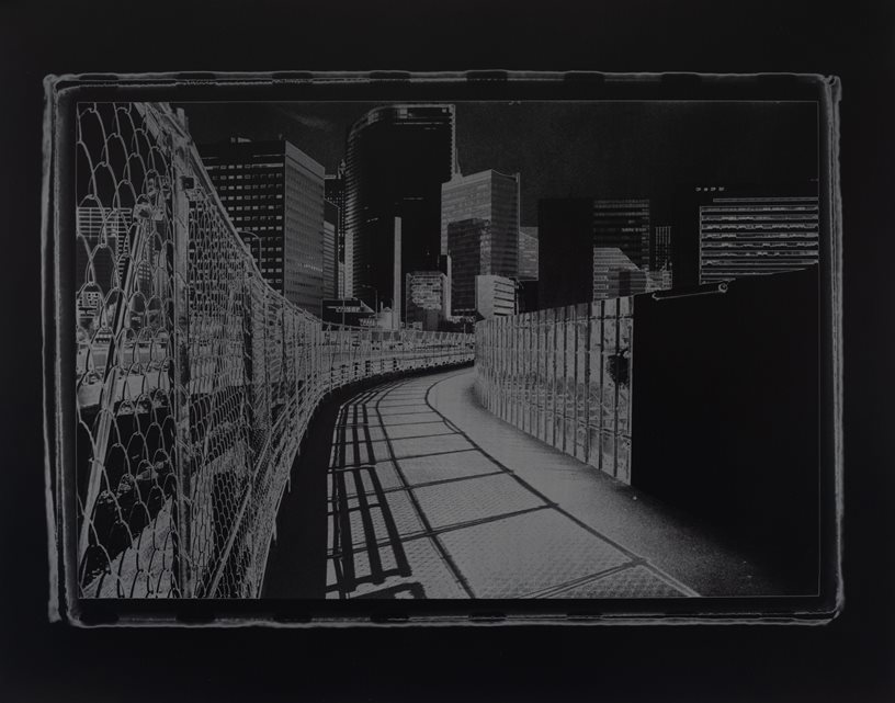 Larry Gawel, Tsukiji No More, clockwise rotation, gelatin silver print, 2023/2024. The image depicts a dark grey image of a right-curved sidewalk through a cityscape with fences on either side of it.