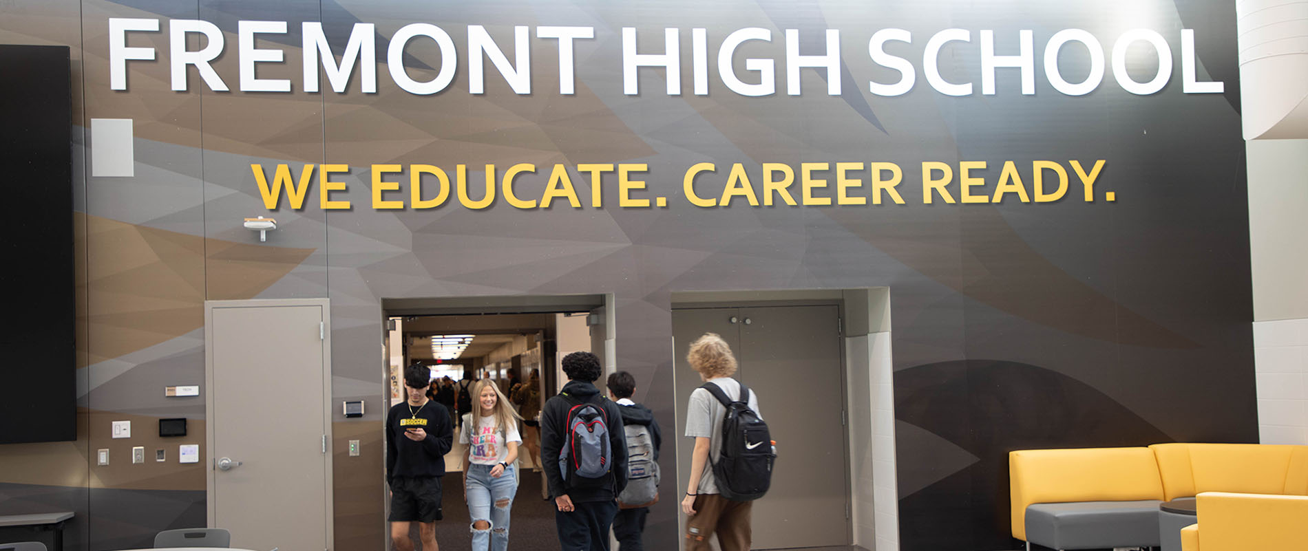 Fremont High School students walk through the hallways during passing period underneath text on the wall in a common area that reads 'Fremont High School. We educate. Career ready.' 