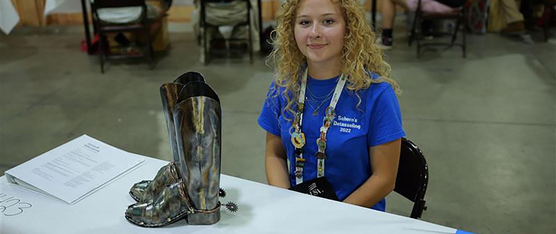Hannah Wieczorek sitting at a table at the SkillsUSA Nebraska competition with welded cowgirl boots she made on display