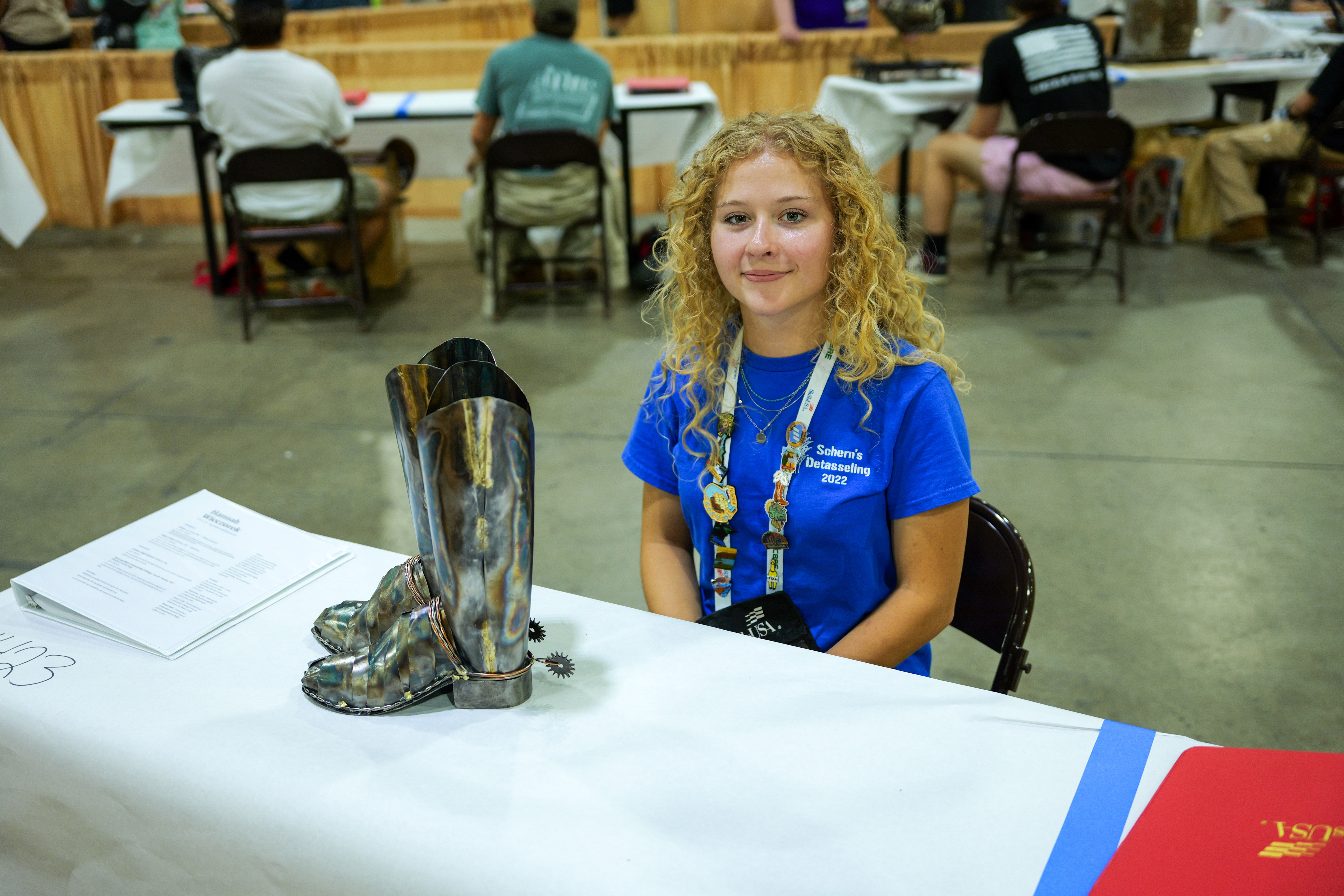 Hannah Wieczorek sitting at a table at the SkillsUSA Nebraska competition with welded cowgirl boots she made on display