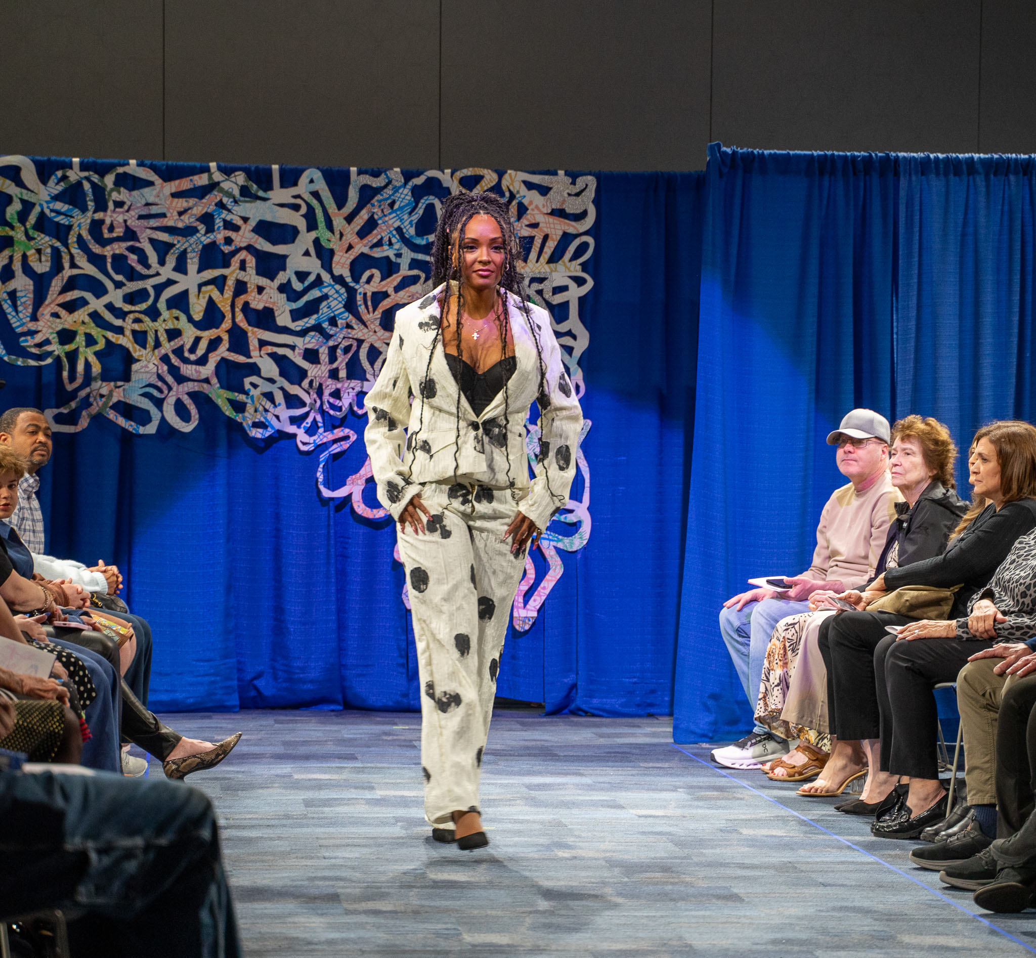 Student model in white suit with black spot walking down runway