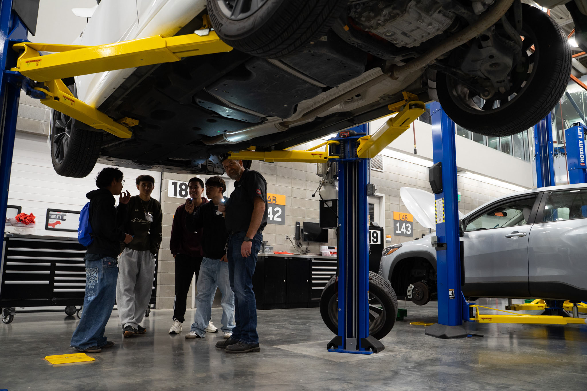 Students and instructors under looking at the underbody of a vehicle while raised on a lift.